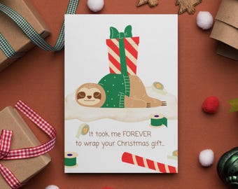 May include: A white card with a cartoon sloth wearing a green sweater and a red and white striped gift box on top of it. The text on the card reads "It took me FOREVER to wrap your Christmas gift."