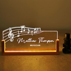 May include: A lighted desk nameplate with a musical theme. The clear acrylic panel features a treble clef, musical notes, and the name "Matthew Thompson, Musician" in white script. The nameplate sits in a wooden base.