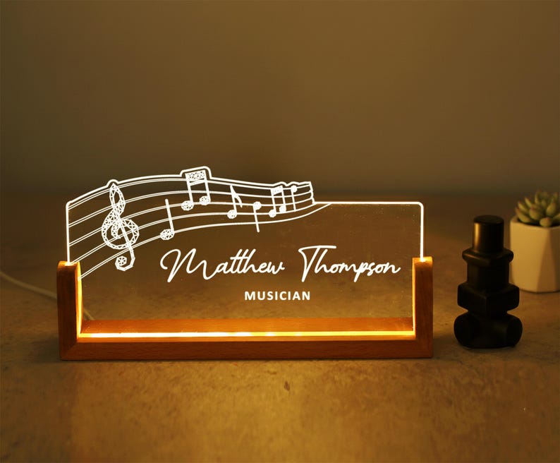 May include: A lighted desk nameplate with a musical theme. The clear acrylic panel features a treble clef, musical notes, and the name "Matthew Thompson, Musician" in white script. The nameplate sits in a wooden base.