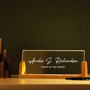 May include: A desk nameplate with the name "Amelia S. Richardson" and the title "CHAIR OF THE BOARD" in elegant script. The nameplate is in a wooden holder, with a pen and a decorative sculpture nearby.