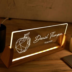 May include: Illuminated desk nameplate with the name "Daniel Thompson" and the word "TEACHER" in white script. A geometric apple design is on the left. The nameplate is set in a wooden base and lit with a warm glow.