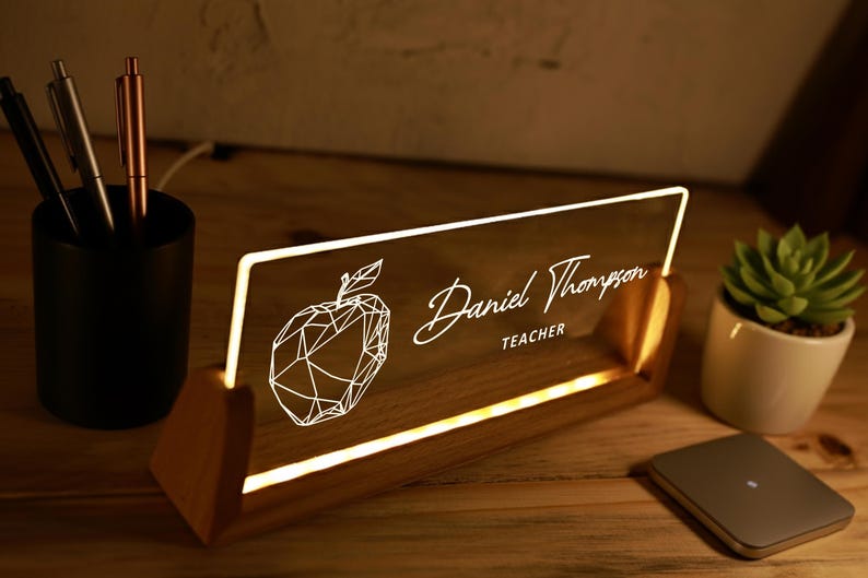 May include: Illuminated desk nameplate with the name "Daniel Thompson" and the word "TEACHER" in white script. A geometric apple design is on the left. The nameplate is set in a wooden base and lit with a warm glow.