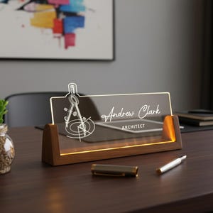 May include: A personalized desk nameplate for an architect. The clear acrylic plate features the name "Andrew Clark" and the title "ARCHITECT" in elegant script. A compass design is etched on the left. The nameplate sits on a wooden base with warm lighting.