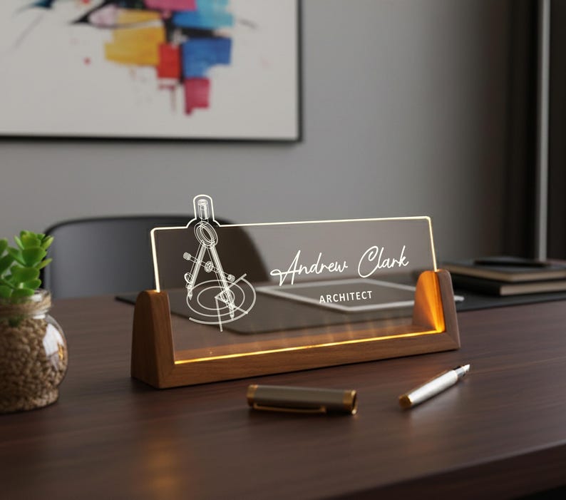 May include: A personalized desk nameplate for an architect. The clear acrylic plate features the name "Andrew Clark" and the title "ARCHITECT" in elegant script. A compass design is etched on the left. The nameplate sits on a wooden base with warm lighting.