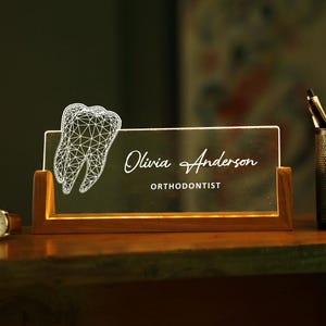 May include: A lighted desk nameplate with a wooden base. The nameplate features the name "Olivia Anderson" and the title "Orthodontist" in elegant script. A wireframe tooth graphic is on the left. A watch and pen holder are also visible.