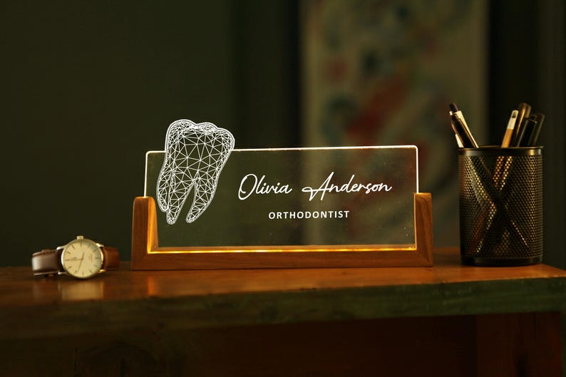 May include: A lighted desk nameplate with a wooden base. The nameplate features the name "Olivia Anderson" and the title "Orthodontist" in elegant script. A wireframe tooth graphic is on the left. A watch and pen holder are also visible.