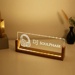 May include: A lighted DJ sign with the text "DJ SOULPHASE" and a headphone graphic. The sign is made of clear acrylic and sits in a wooden base. A gold sculpture and vases are in the background.