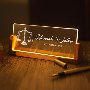 May include: A lighted desk nameplate with a wooden base. The clear acrylic panel displays a scale of justice graphic and the name "Hannah Walker, Attorney at Law" in white script. A pen and pen cap are on the wooden surface.