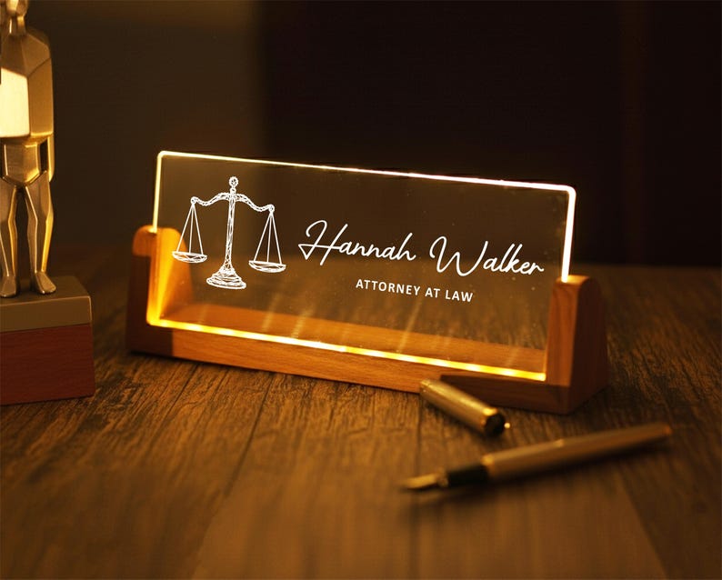 May include: A lighted desk nameplate with a wooden base. The clear acrylic panel displays a scale of justice graphic and the name "Hannah Walker, Attorney at Law" in white script. A pen and pen cap are on the wooden surface.