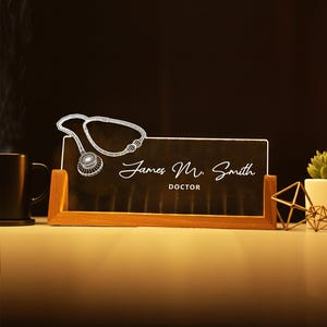 May include: A wooden desk nameplate with a black surface, featuring the name "James M. Smith" and the title "DOCTOR" in white script. A white line drawing of a stethoscope is above the name. The nameplate is set on a wooden base.