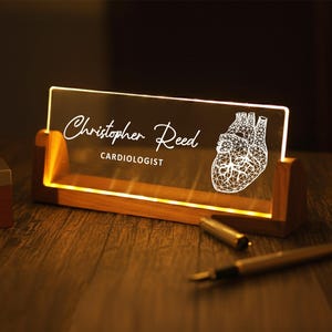 May include: A lighted desk nameplate with the name "Christopher Reed" and the title "CARDIOLOGIST" in white script. A geometric heart design is on the right. The nameplate sits in a wooden base, and a pen rests on the desk.
