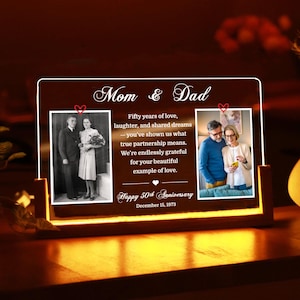 Puede incluir: Un marco de fotos de aniversario personalizado con las palabras "Mom & Dad" en la parte superior. Presenta dos fotos, una en blanco y negro y otra en color, y el texto "Happy 50th Anniversary". El marco está iluminado.