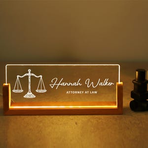 May include: A lighted desk nameplate with a wooden base. The clear acrylic panel displays the name "Hannah Walker" and "ATTORNEY AT LAW" with a scales of justice graphic. A black decorative object and a small potted plant are to the right.