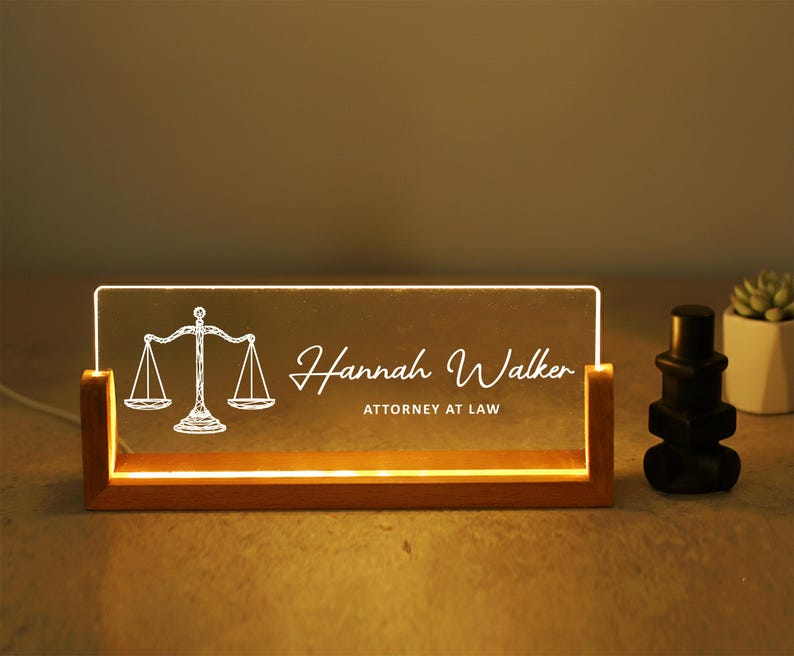 May include: A lighted desk nameplate with a wooden base. The clear acrylic panel displays the name "Hannah Walker" and "ATTORNEY AT LAW" with a scales of justice graphic. A black decorative object and a small potted plant are to the right.