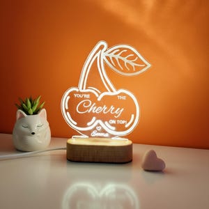 Puede incluir: Lámpara LED con forma de cereza brillante con el texto "You're the Cherry on Top!" y un símbolo de corazón. La lámpara está sobre una base de madera. También se ve una pequeña maceta de cerámica con forma de zorro blanco y un objeto en forma de corazón rosa.