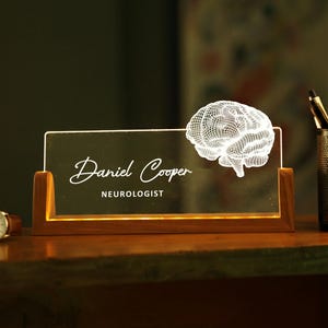 May include: A lighted desk nameplate with the name "Daniel Cooper" and the title "Neurologist" in white script. A 3D brain graphic is on the right. A watch and pen holder are also visible.