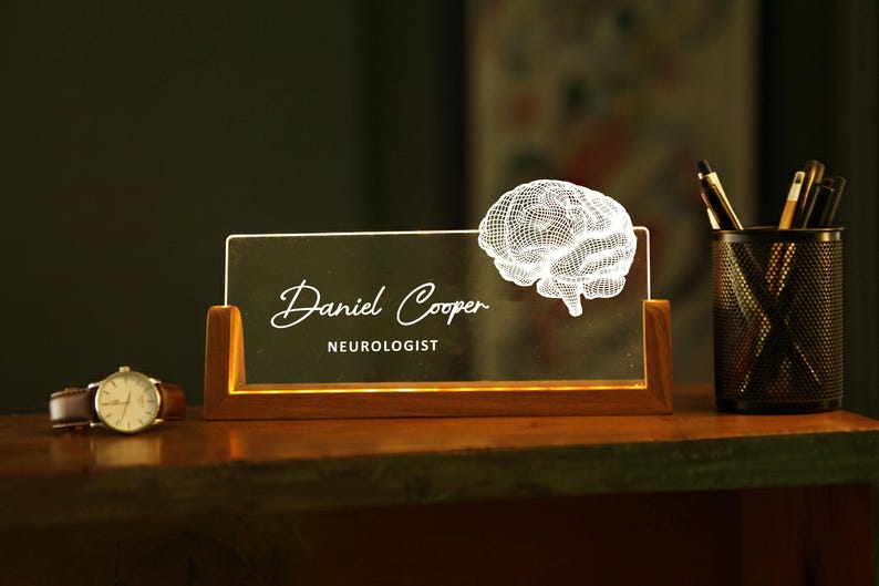 May include: A lighted desk nameplate with the name "Daniel Cooper" and the title "Neurologist" in white script. A 3D brain graphic is on the right. A watch and pen holder are also visible.