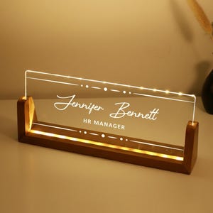May include: A desk nameplate with the name "Jennifer Bennett" and the title "HR MANAGER" etched in white. The clear acrylic plate sits in a wooden base, illuminated by warm white lights. The base is approximately 20 cm long.