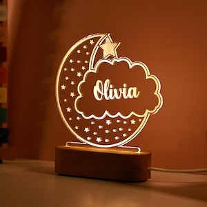 Op de afbeelding: Een houten nachtlampje met een halve maan en wolk ontwerp. De wolk heeft de naam "Olivia" erop geschreven. Het lampje geeft een warm wit licht.