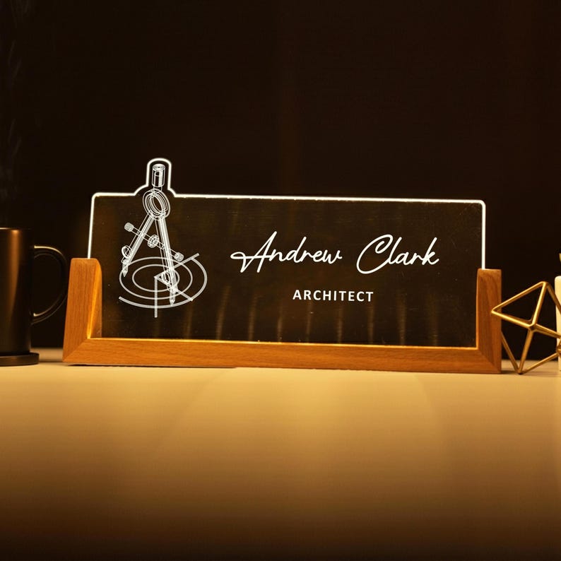 May include: A personalized desk nameplate for an architect. The nameplate features a wooden base and a black surface with the name "Andrew Clark" and the title "ARCHITECT" in white script. A white compass design is on the left.