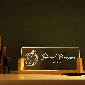 May include: A wooden desk nameplate with the name "Daniel Thompson" and the title "Teacher" in white script. The nameplate features a geometric apple design. A pen rests on the desk.