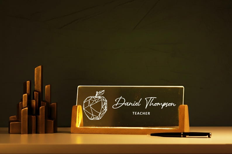 May include: A wooden desk nameplate with the name "Daniel Thompson" and the title "Teacher" in white script. The nameplate features a geometric apple design. A pen rests on the desk.