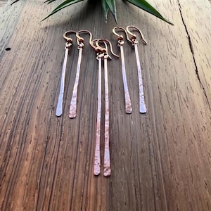 May include: Six pairs of copper-colored, hammered-finish earrings. Each pair features a long, slender bar with a textured lower portion and a simple hook. The earrings are displayed on a wooden surface, with a green plant in the background.