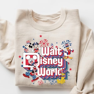 K&ouml;nnte beinhalten: Cremefarbenes Sweatshirt mit einem farbenfrohen Grafikdesign mit Micky und Minnie Maus, Donald Duck und Goofy. Die Grafik zeigt "Walt Disney World" in einem Retro-Schriftzug mit roten, wei&szlig;en und blauen Akzenten und Feuerwerk.
