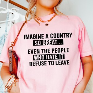 May include: Pink t-shirt with the text "IMAGINE A COUNTRY SO GREAT... EVEN THE PEOPLE WHO HATE IT REFUSE TO LEAVE" in black. The shirt is paired with light blue jeans. A gold necklace and bracelets are visible.