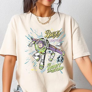 May include: Beige t-shirt featuring a graphic of Buzz Lightyear from Toy Story. The design includes the text "Buzz Lightyear" and "To Infinity and Beyond" in a colorful, cartoon style. The shirt is a casual, short-sleeved style.