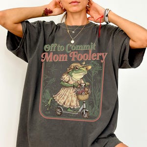 Pode incluir: Camiseta cinza escuro com o texto "Off to Commit Mom Foolery". O gráfico apresenta um sapo com um vestido e chapéu andando de scooter, carregando flores em uma cesta. O design é emoldurado por uma borda vermelha e verde.