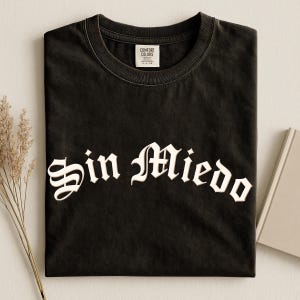 May include: Black t-shirt featuring the white gothic text "Sin Miedo". The shirt is neatly folded, with a label visible at the collar. A sprig of dried grass and a book are in the background.