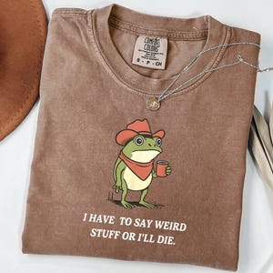 Puede incluir: Camiseta Comfort Colors marrón con una rana de dibujos animados con sombrero de vaquero y taza. El texto "I HAVE TO SAY WEIRD STUFF OR I'LL DIE" está impreso debajo. Un collar plateado está sobre la camiseta.