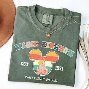 K&ouml;nnte beinhalten: Ein olivgr&uuml;nes T-Shirt mit einer farbenfrohen Grafik mit den Worten "MAGIC KINGDOM" und einer Mickey Mouse-Silhouette mit einem Schlossdesign. Das Shirt enth&auml;lt auch den Text "EST 1971" und "WALT DISNEY WORLD". Eine goldene Halskette liegt auf dem Shirt.