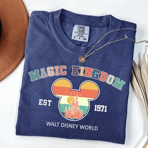 K&ouml;nnte beinhalten: Ein marineblaues T-Shirt mit den Worten "MAGIC KINGDOM" in bunten Buchstaben &uuml;ber einem Mickey Mouse-Kopf-Design mit einem Schloss und Sonnenuntergang. Das Shirt hat auch den Text "EST 1971" und "WALT DISNEY WORLD". Eine goldene Halskette liegt &uuml;ber dem Shirt.