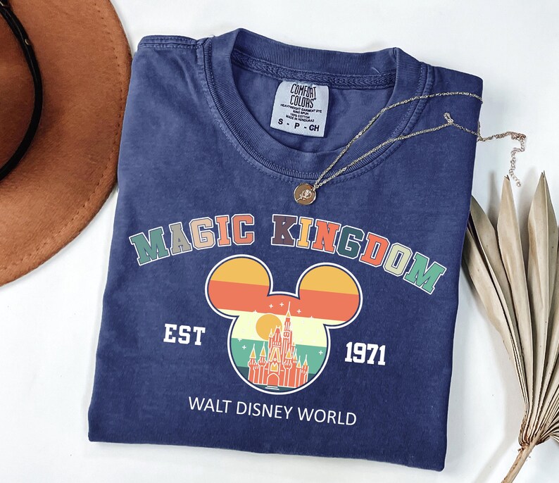 K&ouml;nnte beinhalten: Ein marineblaues T-Shirt mit den Worten "MAGIC KINGDOM" in bunten Buchstaben &uuml;ber einem Mickey Mouse-Kopf-Design mit einem Schloss und Sonnenuntergang. Das Shirt hat auch den Text "EST 1971" und "WALT DISNEY WORLD". Eine goldene Halskette liegt &uuml;ber dem Shirt.