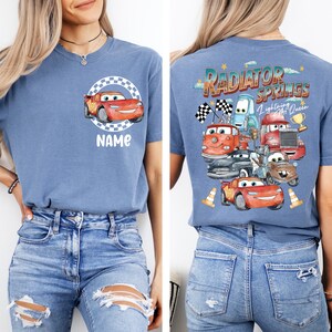 May include: Blue t-shirt featuring a cartoon car design with the text "NAME" on the front. The back of the shirt displays a graphic of various cartoon cars with the text "RADIATOR SPRINGS Lightning McQueen".