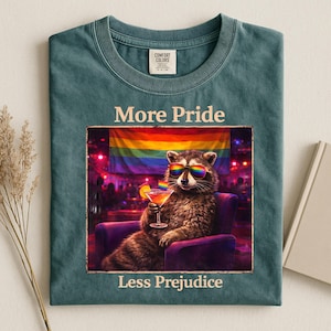 Op de afbeelding: Teal Comfort Colors T-shirt met de tekst "More Pride Less Prejudice". Het shirt toont een wasbeer met een regenboog zonnebril, die een cocktail vasthoudt en in een paarse stoel zit voor een regenboogvlag.