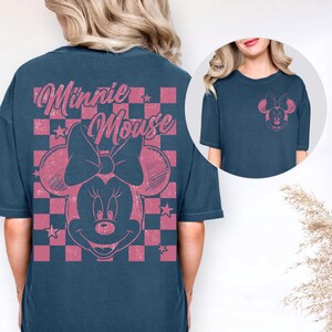 May include: A navy blue t-shirt featuring a pink Minnie Mouse graphic on the front and back. The back design includes the words "Minnie Mouse" above the character's face, set against a pink and black checkered background. The front has a smaller Minnie Mouse graphic.