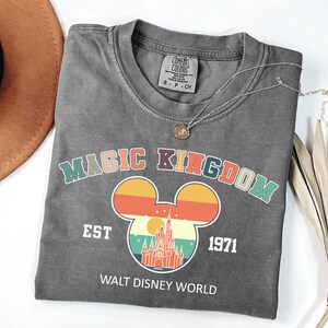 K&ouml;nnte beinhalten: Ein graues T-Shirt mit den Worten "MAGIC KINGDOM" in bunten Buchstaben und einem Mickey Mouse Kopf-Design mit einem Schloss darin. Das Shirt hat auch den Text "EST 1971" und "WALT DISNEY WORLD". Eine goldene Halskette liegt &uuml;ber dem Shirt.