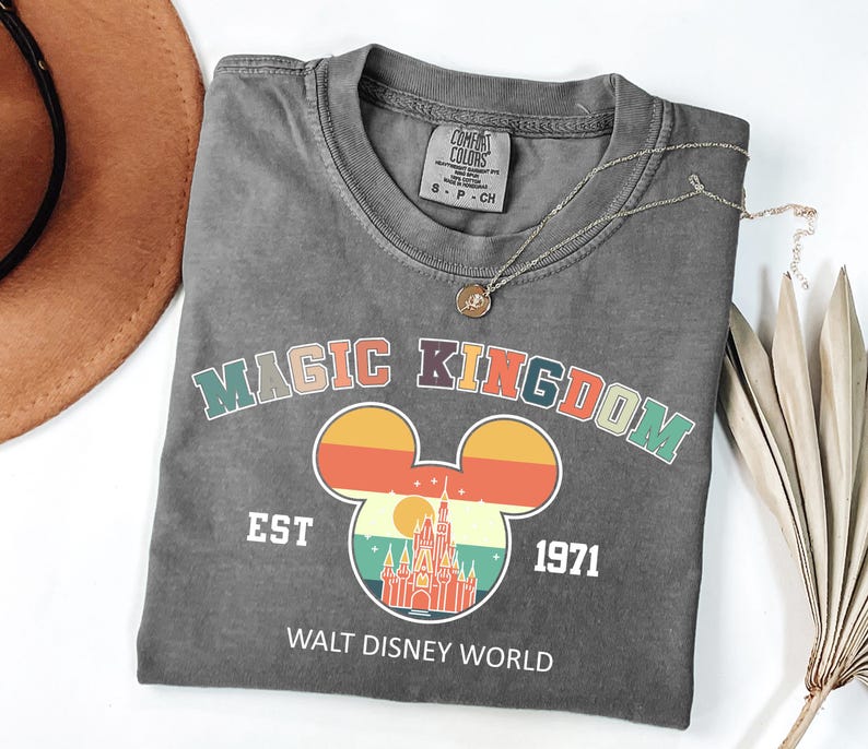 K&ouml;nnte beinhalten: Ein graues T-Shirt mit den Worten "MAGIC KINGDOM" in bunten Buchstaben und einem Mickey Mouse Kopf-Design mit einem Schloss darin. Das Shirt hat auch den Text "EST 1971" und "WALT DISNEY WORLD". Eine goldene Halskette liegt &uuml;ber dem Shirt.