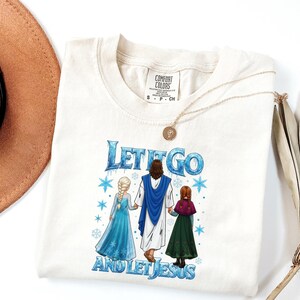 May include: Cream-coloured t-shirt with the text "LET IT GO AND LET JESUS" in blue. The graphic features Jesus holding hands with two cartoon characters. A gold necklace with a pendant rests on the shirt. The shirt is a Comfort Colors brand.