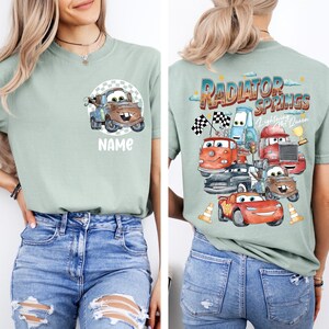 May include: Sage green t-shirt featuring a cartoon image of Mater from the movie Cars, with the word "NAME" below. The back of the shirt displays a graphic of various Cars characters with the text "Radiator Springs Lightning McQueen".