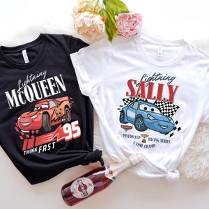 May include: Two t-shirts featuring Lightning McQueen and Sally Carrera from the Cars movie. One black shirt has McQueen with the text "Lightning McQueen 95 Think Fast". The other is white with Sally and the text "Lightning Sally".