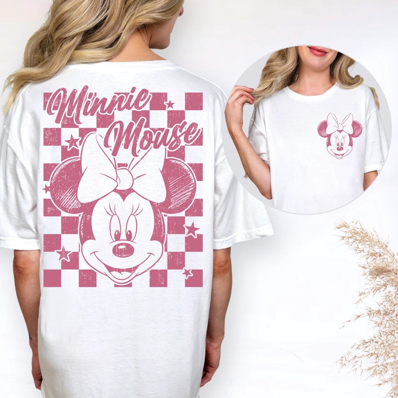 May include: White t-shirt featuring a pink Minnie Mouse graphic. The back of the shirt has a large Minnie Mouse design with the words "Minnie Mouse" in a script font over a pink and white checkered pattern. A smaller Minnie Mouse graphic is on the front.