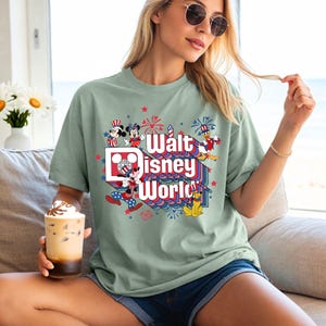 K&ouml;nnte beinhalten: Salbeigr&uuml;nes T-Shirt mit einer farbenfrohen Grafik von Disney-Figuren und dem Schriftzug "Walt Disney World". Das Design umfasst Mickey, Minnie, Donald und Pluto, mit Feuerwerk und patriotischen Sternen.