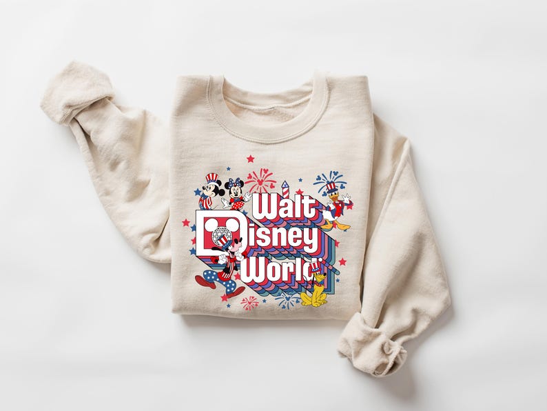 K&ouml;nnte beinhalten: Cremefarbenes Sweatshirt mit einem farbenfrohen Grafikdesign mit Micky und Minnie Maus, Donald Duck und Goofy. Die Grafik zeigt "Walt Disney World" in einem Retro-Schriftzug mit roten, wei&szlig;en und blauen Akzenten und Feuerwerk.