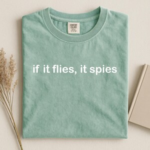 Puede incluir: Camiseta verde claro con la frase blanca "if it flies, it spies". La camiseta tiene cuello redondo y una etiqueta en el cuello. La prenda est&aacute; doblada y descansa sobre una superficie neutra.