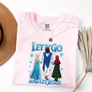 May include: A light pink Comfort Colors t-shirt with the text "Let It Go and Let Jesus" in blue and white. The graphic features three figures in a winter scene. A gold necklace with a pendant is on the shirt.