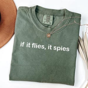 Puede incluir: Camiseta verde oliva con la frase blanca "if it flies, it spies". Un collar dorado con un peque&ntilde;o colgante descansa sobre la camiseta. La camiseta est&aacute; doblada sobre una superficie blanca.